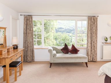 A room with a chaise lounge and pillows in front of a large window and a wooden dressing table with a lamp and stool at Huntingstile Lodge in Grasmere