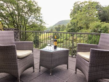 A patio with two wicker chairs and a wicker table holding a wine bottle and two glasses overlooking trees and hills at Huntingstile Lodge in Grasmere