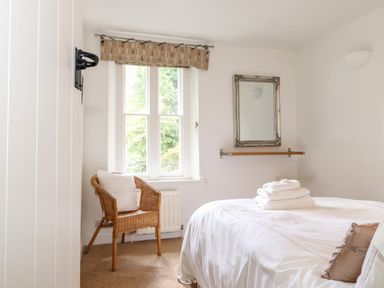 A bedroom with a bed, wicker chair with cushion, window, wall mirror, and folded towels on the bed at Huntingstile Lodge in Grasmere