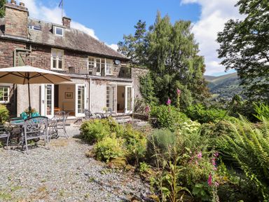 A garden patio with tables, chairs, and an umbrella outside a stone house with open doors and a balcony at Huntingstile Lodge in Grasmere