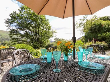 An outdoor dining table with turquoise plates and glasses under a beige umbrella surrounded by greenery and trees at Huntingstile Lodge Grasmere