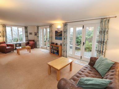 A living room with brown leather sofa and chairs wooden tables bookshelf and large windows with curtains at Huntingstile Lodge in Grasmere