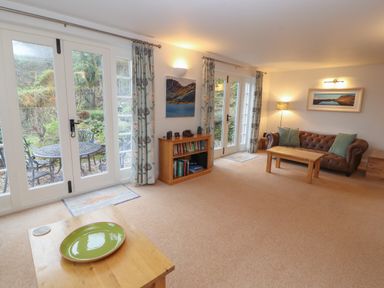 A living room with brown leather sofa green cushions wooden tables a bookshelf and glass doors leading to a garden at Huntingstile Lodge in Grasmere