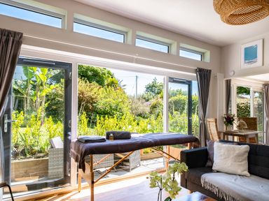 A living room with a massage table near large windows overlooking a garden at Chi Lowarth in Porthleven