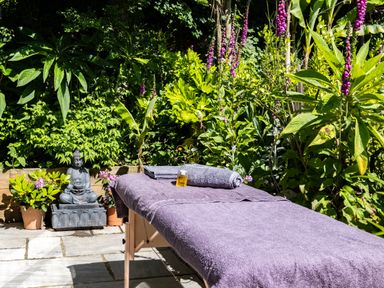 An outdoor massage table with a towel and oil bottle surrounded by plants and a Buddha statue at Chi Lowarth in Porthleven