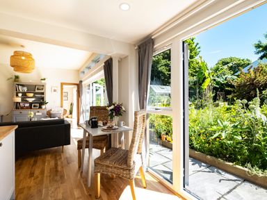 A dining table with two chairs next to large open glass doors leading to a garden with plants and trees at Chi Lowarth in Porthleven