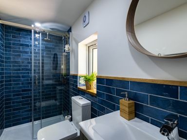 A bathroom with a glass shower enclosure blue tiled walls a toilet a sink with a soap dispenser and a round mirror at Chi Lowarth in Porthleven
