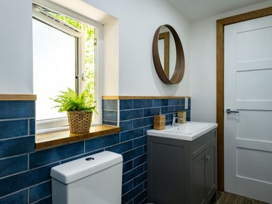 A bathroom with a toilet a window with a plant a sink with soap dispensers a round mirror and wooden door frame at Chi Lowarth in Porthleven