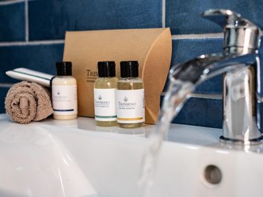 A bathroom sink with running water and toiletries on the counter including shampoo shower gel lotion and a rolled towel at Chi Lowarth in Porthleven