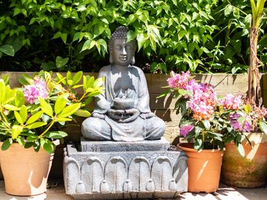 A garden scene with a Buddha statue water fountain surrounded by potted plants with pink flowers at Chi Lowarth in Porthleven
