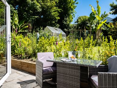 An outdoor patio area with a glass table and wicker chairs overlooking a garden with a greenhouse and various plants at Chi Lowarth in Porthleven