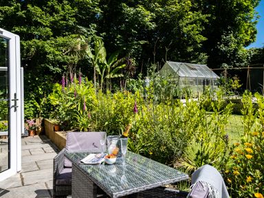 A patio with a glass table and chairs next to a garden with plants and a greenhouse at Chi Lowarth in Porthleven
