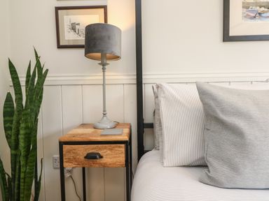 A bedside table with a lamp and a coaster next to a bed with pillows and a plant in the corner at Chi Lowarth in Porthleven