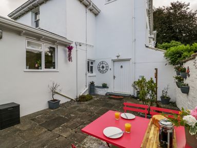An outdoor patio with a white door and clock on the wall red table and chairs with plates glasses of orange juice and a coffee press at Pentre Court Cottage in Abergavenny