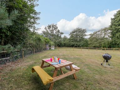 An outdoor picnic area with a wooden table set with plates and cups a barbecue grill and a fenced grassy field at Pentre Court Cottage in Abergavenny