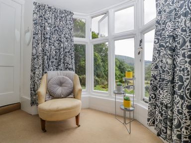 A corner with a beige armchair a round cushion three small potted plants on a stand and large windows with patterned curtains at Pentre Court Cottage in Abergavenny