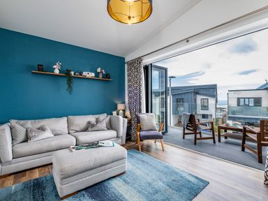 A living room with a grey sofa and ottoman next to a glass door leading to a balcony with chairs at Beach Haven in Perranporth