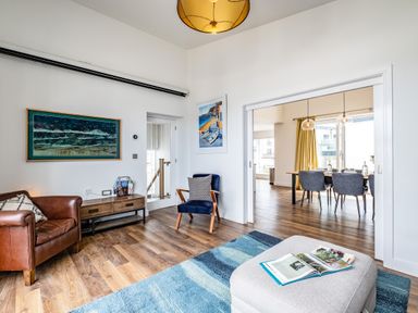 A living room with a brown leather armchair a blue chair a wooden side table and a gray ottoman with an open book at Beach Haven in Perranporth