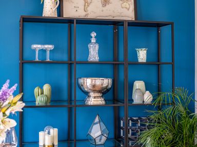 A black metal shelving unit against a blue wall with glassware, decorative bowls, vases, candles, a small plant, and a world map at Beach Haven in Perranporth