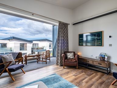 A living room with wooden chairs a leather armchair a wooden sideboard and a large window showing a balcony at Beach Haven in Perranporth