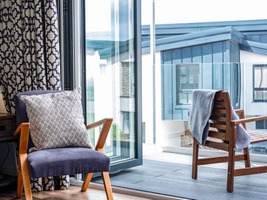 A chair with a patterned pillow next to open glass doors leading to a balcony with a wooden chair and a draped blanket at Beach Haven in Perranporth
