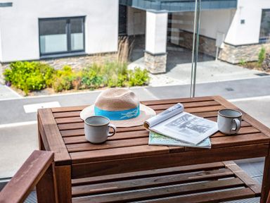 A wooden table on a balcony with two mugs a hat and an open magazine with a building and plants visible outside