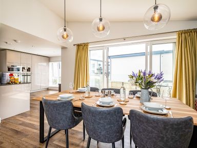 A dining area with a wooden table set for six with plates glasses and a flower vase next to a kitchen with built in appliances at Beach Haven in Perranporth