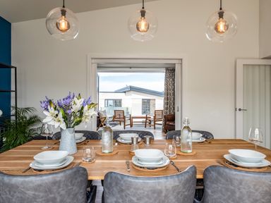 A dining table set with plates glasses cutlery and a vase of flowers in a dining room with pendant lights and an open door leading to a balcony with chairs at Beach Haven in Perranporth