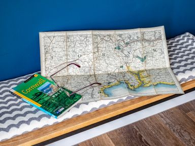 A folded map and a Cornwall walking guidebook with glasses on a bench with a patterned blanket at Beach Haven in Perranporth