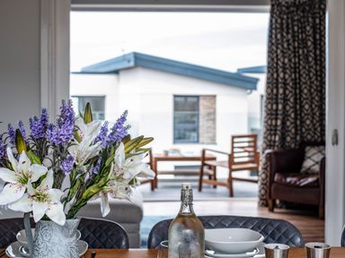 A dining table with a vase of flowers and set plates in a room with a view of a balcony with wooden chairs at Beach Haven in Perranporth
