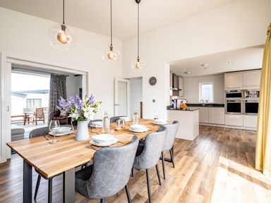 A dining area with a wooden table set with plates and glasses next to a kitchen with built in appliances at Beach Haven in Perranporth
