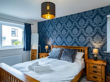 A bedroom with a wooden bed frame, white bed linens, gray pillows, bedside tables with lamps and a full length mirror at Beach Haven in Perranporth