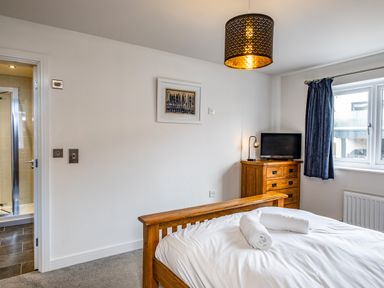 A bedroom with a wooden bed and white bedding with rolled towels on it a wooden dresser with a small lamp and television a window with blue curtains a door leading to a bathroom with a glass shower at Beach Haven in Perranporth