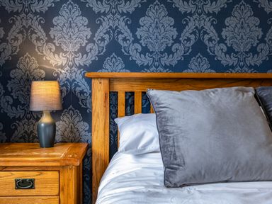 A wooden bed with large gray pillows and a wooden nightstand with a lamp against patterned wallpaper at Beach Haven in Perranporth