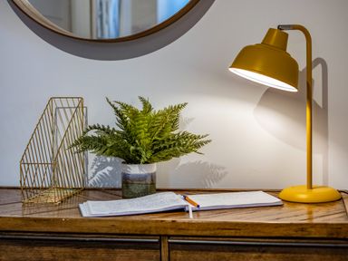 A wooden table with a yellow desk lamp a green plant in a pot an open notebook with a pen and a gold wire organizer at Beach Haven in Perranporth
