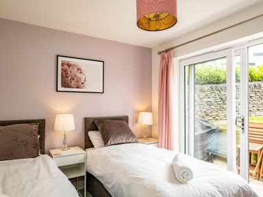A bedroom with two single beds side by side with white bedding and brown pillows with bedside tables with lamps and a window with pink curtains at Beach Haven in Perranporth