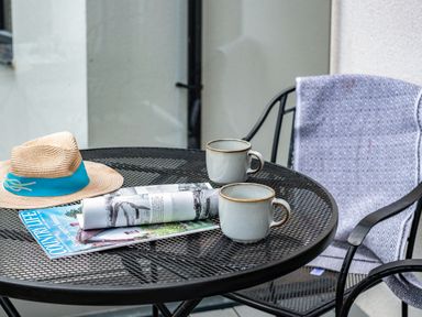 A metal outdoor table with two mugs a magazine a rolled magazine and a hat next to a chair with a blanket Beach Haven in Perranporth