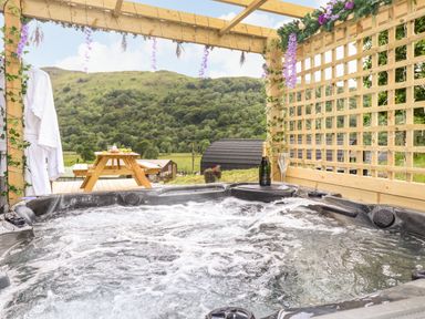An outdoor hot tub under a wooden pergola with hanging flowers and a bottle with glasses next to it and a picnic table in the background at Hartsop Magic - Crossgate Luxury Glamping in Hartsop near Glenridding