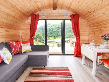 An interior of a wooden cabin with a gray sofa red cushions a striped rug a white dining table with chairs and red curtains at Hartsop Magic - Crossgate Luxury Glamping in Hartsop near Glenridding