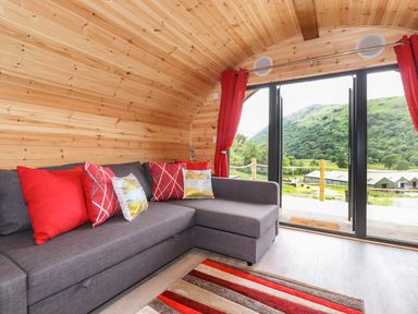 A wooden room with a grey sectional sofa and red cushions next to glass doors overlooking green hills at Hartsop Magic - Crossgate Luxury Glamping in Hartsop near Glenridding