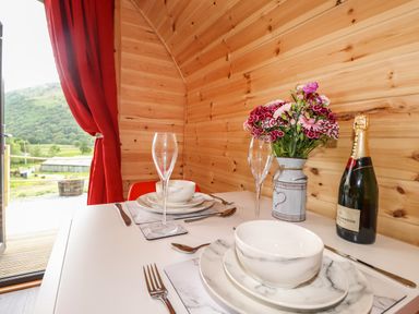A small dining table set with plates bowls cutlery glasses a bottle of champagne and a flower vase in a wooden cabin with red curtains at Hartsop Magic - Crossgate Luxury Glamping in Hartsop near Glenridding