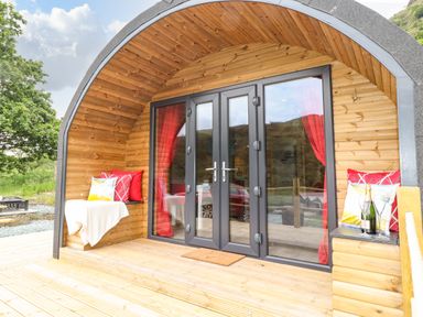 An arched wooden cabin with glass double doors red curtains and cushioned seating on the porch at Hartsop Magic - Crossgate Luxury Glamping Hartsop near Glenridding