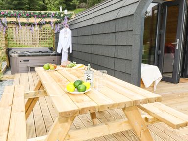 An outdoor wooden picnic table with fruit and a bottle next to a hot tub and a robe hanging on a pergola at Hartsop Magic - Crossgate Luxury Glamping in Hartsop near Glenridding