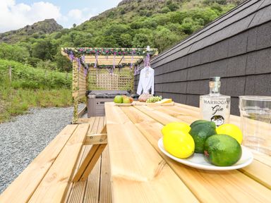 An outdoor wooden picnic table with fruit and a bottle of gin near a hot tub under a pergola decorated with flowers at Hartsop Magic - Crossgate Luxury Glamping in Hartsop near Glenridding