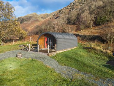 A small cabin with a curved roof next to a picnic table on grass with hills and trees in the background at Hartsop Magic - Crossgate Luxury Glamping in Hartsop near Glenridding