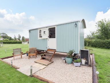 A small light blue wooden cabin with white door and window on a gravel area with two wooden chairs and potted plants at Honeybee Lodge in Stixwould near Woodhall Spa