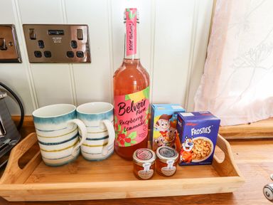A wooden tray with two mugs, a bottle of raspberry lemonade, two small jars of orange marmalade, and two cereal boxes on a kitchen counter at Honeybee Lodge in Stixwould near Woodhall Spa