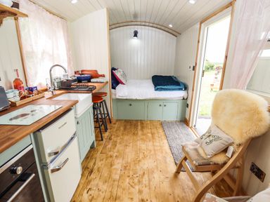 A compact room with a small kitchen area including a sink and refrigerator a bed with storage underneath a wooden chair with a cushion and fur throw and an open door showing outdoor greenery at Honeybee Lodge in Stixwould near Woodhall Spa