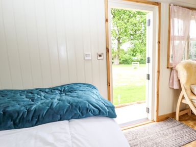 A bedroom with a bed covered in a blue comforter and white sheets with an open door showing a green lawn and trees at Honeybee Lodge in Stixwould near Woodhall Spa