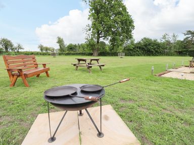 A grassy outdoor area with a fire pit and cooking pans wooden bench and picnic table surrounded by trees at Honeybee Lodge in Stixwould near Woodhall Spa